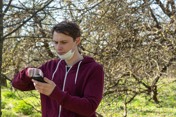 young man browses web pages on a mobile device. the action takes place in the forest. on the chin is a white medical protective mask. Protection against coronavirus, viruses and diseases.