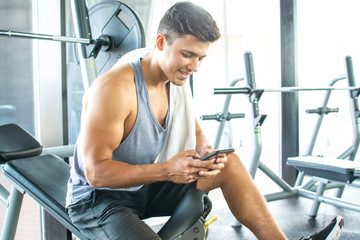 Handsome young man resting on weight bench and using phone after training at gym.