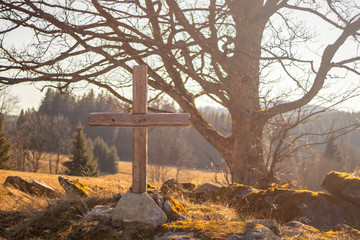 wooden cross on stones, in the background a big old tree, meadow and forests, in the evening