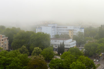 Fototapeta na wymiar Lowrise buildings and streets with green trees in dense fog at summer day. Thick fog came from the sea and covered the city. Climate and weather changes.