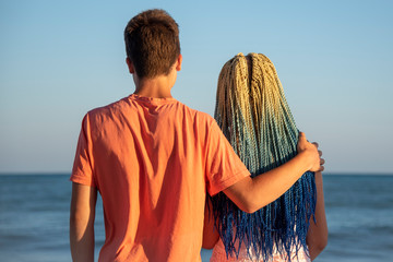 Two teenagers embracing stand with his back to the camera and look at the waves crashing on the rocks.The spray flies in all directions.The girl with the blue Senegalese braids.