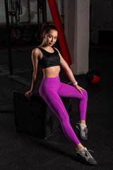 Beautiful young caucasian girl posing in a dark gym. A woman in sportswear is resting from a circular workout on a box. A break between sets.