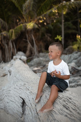 Happy smile european blond baby boy have rest and sit on white sand beach. . white cotton clothes. t-short.Thailand. palms. rocks