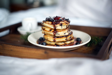Delicious stack of blueberry pancakes with syrup poured over the blueberries dripping onto the pancakes is served in bed on a small tray with an espresso