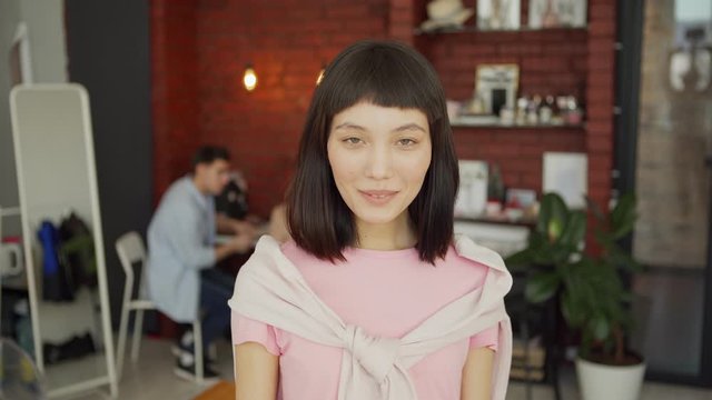 Panning medium shot portrait of pretty young Asian woman with sweater over her shoulders looking at camera and smiling standing in modern loft office