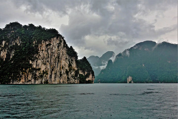 A foggy and rainy day on the unique Cheo Lan Lake. Rocks covered with green plants rise steeply from the water. On their tops are shreds of clouds. Quiet and mysterious.