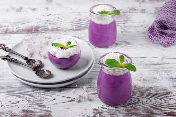 Bilberry mousse topped with whipped cream and mint in glass jars on the white wooden table