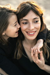 Loving mother and daughter gently hug each other on the beach at sunset, they smile and enjoy communication. A photo with a blurred background.