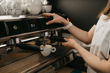 A female barista with white apron brewing espresso in a professional espresso machine in a coffee shop. A close-up photo of a coffee pouring in a white cup from a coffee machine in a cafe.