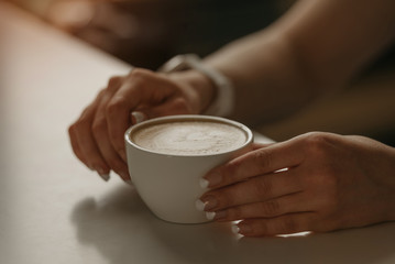 A girl with beautiful nails keeps a cup of latte in her hands in a cafe.