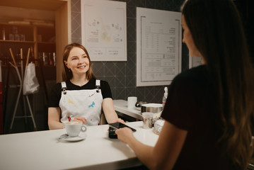 A female barista smiling holds out to a client a terminal for paying for a cup of coffee. A woman with long hair paying for a latte with a smartphone by contactless NFC technology in a cafe.