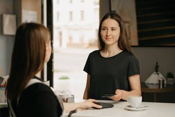 A happy girl with long hair paying for her coffee with a smartphone by contactless NFC technology in a cafe. A brunette female barista holds out to a client a terminal for paying.