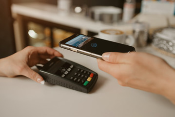 A woman paying for her latte with a smartphone by contactless PAY PASS technology in a cafe. A female barista holds out a terminal for paying to a client in a coffee shop.