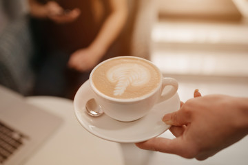 A close-up photo of a latte which a female barista holds out to a girl in a coffee shop. A young woman working remotely on a laptop waits for a cup of coffee in a cafe.