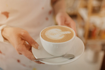 A close-up photo of a latte which a female barista holds in her hands in a coffee shop. A barista preparing an order in a cafe.