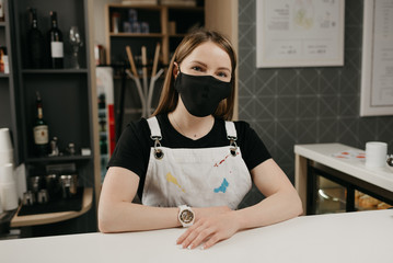 A happy female barista in a medical black face mask smiles and waiting for clients in the coffee shop. A beautiful female owner of the cafe poses with arms crossed behind the bar.