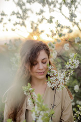Fototapeta premium A young girl with long hair in a beige trench coat and white dress next to a flowering tree, girl's face in flowers