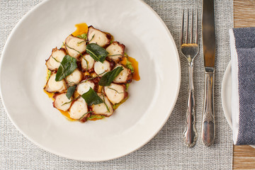 Charred grilled Octopus with oil and Smoked salad leaves on a plate on a table with fork and knife, from above. Grilled Octopus with lemon and tomato, Mykonos, Greece
