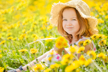 little cute girl 3 years old smiling in a straw hat sits on the grass in yellow dandelions in the summer with a place for text