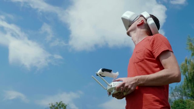 Man controls a quadrocopter through a remote control and looks at the video with goggles on his head. Drone operator in red T-shirt in a park shoots video on drone. Blue sky on the background