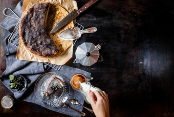 Apple cinnamon cake on a dark table. Coffee pot, a cup of coffee, milk on a dark background. Female hand pours milk into coffee. Space for text.