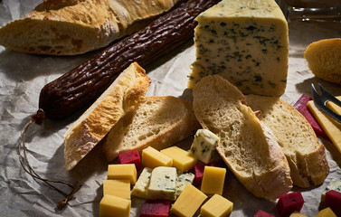 Cheese plate of different kinds and pieces on parchment, with special knife and sliced fresh baguette, with stick of dry sausage, wine glasses on dark wooden background.