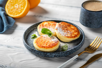 Cottage cheese pancakes on a plate, white wooden table, close-up. Delicious and beautiful breakfast with coffee.