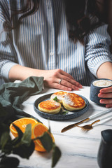 Woman at the table with a beautiful and tasty breakfast. Cottage cheese pancakes on a plate, cup with coffee and oranges on a white wooden background.