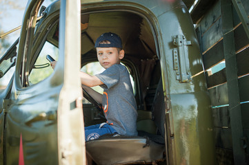 A little boy in a cap sits behind the wheel of a large truck, the door is open, looks away