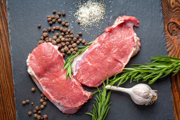 Raw meat and spices. Top view of two slices of fresh raw meat, garlic, rosemary and spices on a black slate stone plate. Preparing meat for a barbecue.