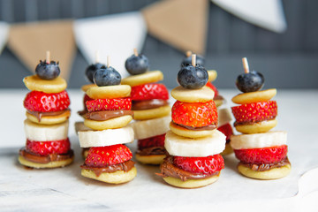 Mini pancakes canape with berries, chocolate cream and banana, new trendy social media food. Party healthy dessert bar on white table. Close up view, copy space. Small pancakes with fresh fruits.