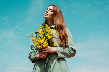 Portrait of redheaded young woman with eyes closed standing against sky holding bunch of yellow flowers