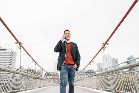 Smiling businessman talking on smart phone while walking on bridge against sky in Frankfurt, Germany