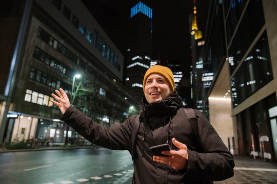 Portrait of smiling man with smartphone haling taxi at night, Frankfurt, Germany