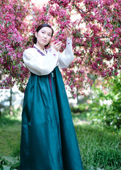 A young Korean girl in a beautiful white and sea-coloured traditional costume hanbok surrounded by blossoming pink cherry trees poses on camera