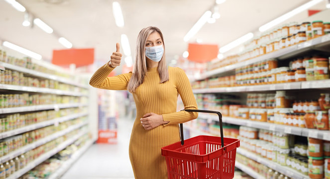 Woman with a shopping basket wearing a protective face mask inside a supermarket and showing thumbs up