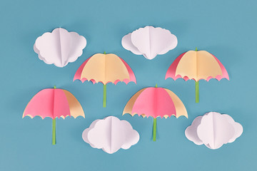 Rainy weather concept with paper craft clouds and pink and yellow umbrellas on blue background