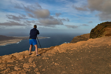 Man taking photograph from dramatic clifftop location near Mirador del Rio in Lanzarote, Spain. Man has tripod. Taken from behind, not identifiable.