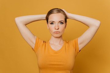 Beautiful young red haired woman in orange t-shirt, collects her hair on the head by both hands, looks confident, over yellow background. Purposeful people concept.