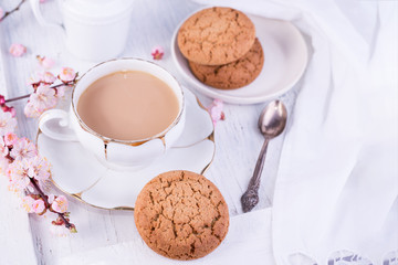 White porcelain cup of coffee or cacao, milk jug, and fresh-baked oat cookies. English breakfast still-life with drink and treats and tablecloth.