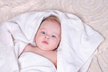 Newborn baby with light hair and blue eyes lies on bed on light blanket in white, soft, dry towel after bathing procedure. Concept of baby's daily routine and child hygiene