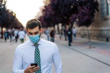 Young elegant businessman stands in middle of crowded street with surgical mask looking a phone