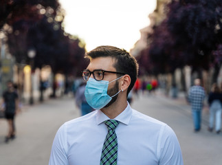 Young elegant businessman stands in middle of crowded street with surgical mask