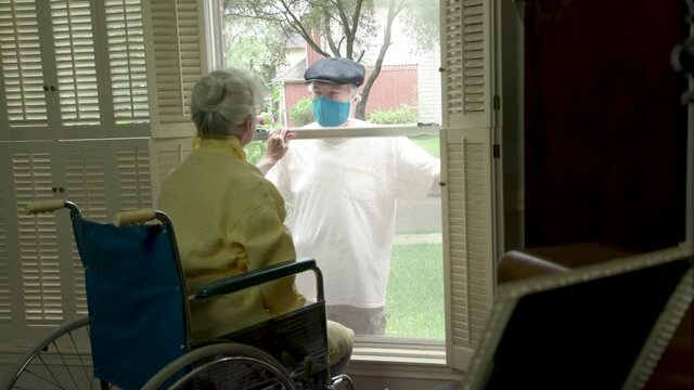 An Elderly Woman In A Wheelchair Social Distancing Because Of COVID19 Visits With A Caring Relative Or Neighbor Through A Window.