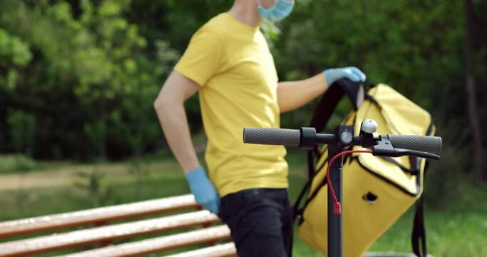 A mask and gloves courier receives an order for food delivery, puts on a thermal backpack and leaves the camera frame.