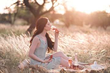 Obraz premium Stylish woman drinking wine with strawberry siting in field with picnic outdoor at sunset. French style. 20s. Holiday season.