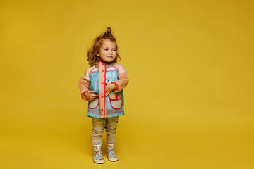 Stylish little girl in modish raincoat isolated at the yellow background. Child fashion. Copy space