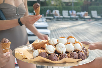 Waiter with metal full tray offer variety of many vanilla and chocolate cone waffle ice cream dessert at pool party celebration event near beach poolside at summertime day. Catering food service