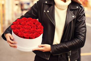 Outdoor portrait of young beautiful woman holding white box with red roses, wearing leather jacket - Powered by Adobe