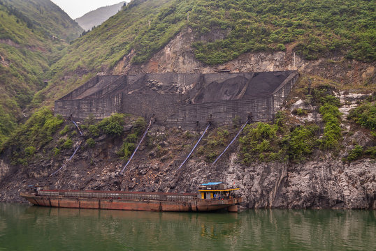 Guandukou, Hubei, China - May 7, 2010: Wu Gorge in Yangtze River. wall structure above coal loading dock on shoreline of green foliage covered mountain  with barge present on green water.